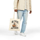 A person wearing a cream button-up shirt, light blue jeans, and white sneakers holds a cream canvas tote bag with a Halloween graphic that reads 'NO TRICKS JUST TREATS' with a ghost and pumpkin illustration, against a plain white background.