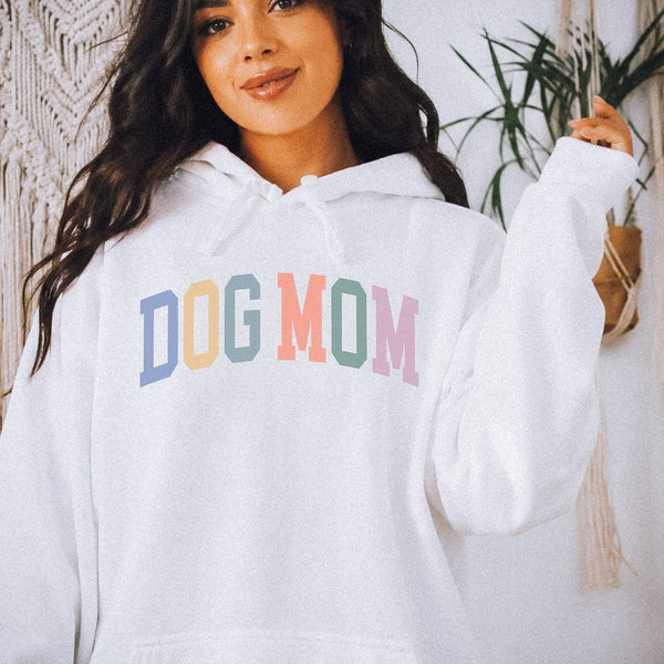 A woman wearing a white pullover hoodie with the words 'DOG MOM' printed across the chest in pastel blue, peach, yellow, green, purple and pink letters. She is posing in a cozy indoor setting with boho decor.