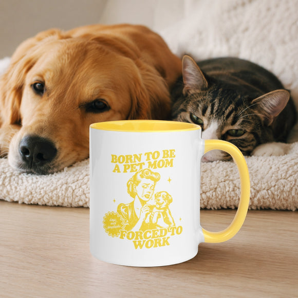 A white ceramic mug with a yellow handle and interior sits on a wooden surface in front of a golden retriever and a tabby cat resting on a cream blanket. The mug is printed with a yellow graphic and the text 'BORN TO BE A PET MOM FORCED TO WORK'.