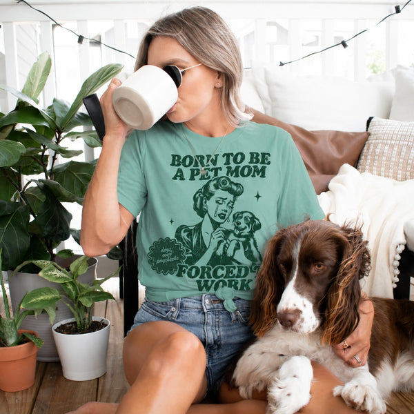 A woman wearing a seafoam green graphic t-shirt with a dog illustration and the text 'BORN TO BE A PET MOM FORCED BY WORK' sits on a couch with her brown and white dog, while drinking from a white mug.