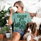 A woman wearing a seafoam green graphic t-shirt with a dog illustration and the text 'BORN TO BE A PET MOM FORCED BY WORK' sits on a couch with her brown and white dog, while drinking from a white mug.