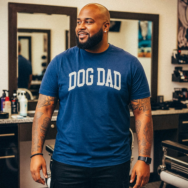 A man with a shaved head and beard wearing a blue crewneck t-shirt with 'DOG DAD' printed in white across the chest, standing in a barbershop environment.