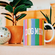 A ceramic mug with vertical rainbow stripes and the words 'DOG MOM' in white, styled on a tabletop with potted plants and a disco ball in the background.