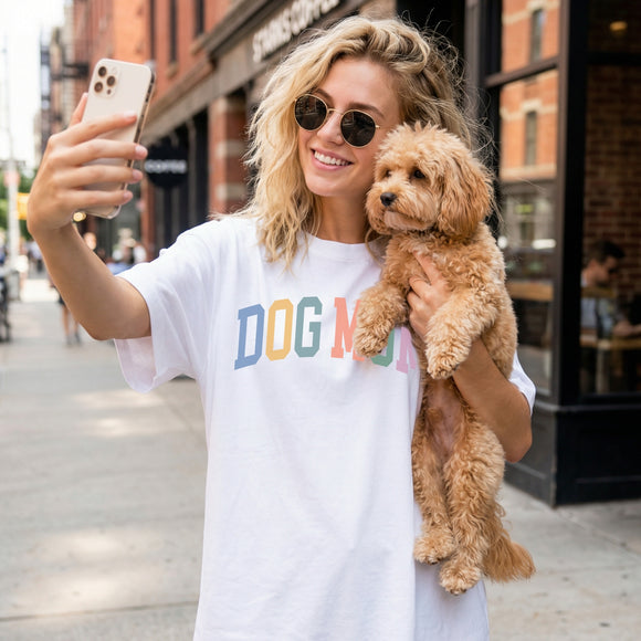 A woman taking a selfie on a city street while holding a small fluffy dog. She is wearing a white graphic T-shirt with the text 'DOG MOM' printed across the chest in blue, orange, and green letters.