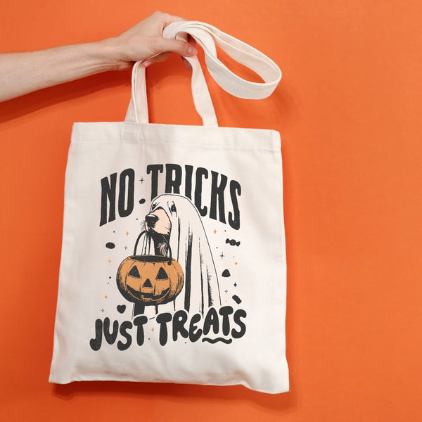 An off-white canvas tote bag with a Halloween graphic print reading 'NO TRICKS JUST TREATS' and showing a ghost, pumpkin, and dog, held by a hand against a solid orange background.